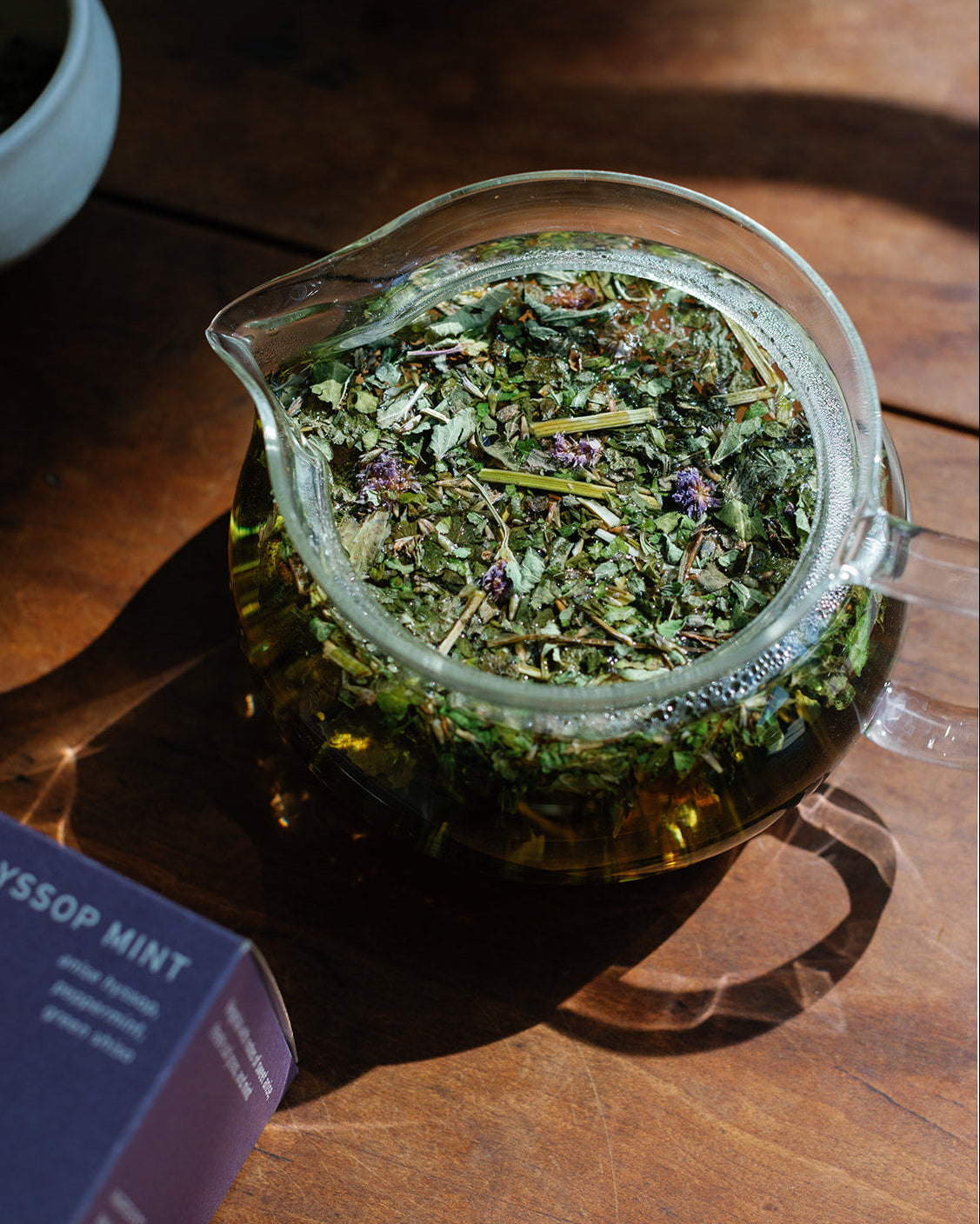 Looseleaf herbal tea in a glass teapot on a wooden surface with a box labeled 'Hyssop Mint' in the background.