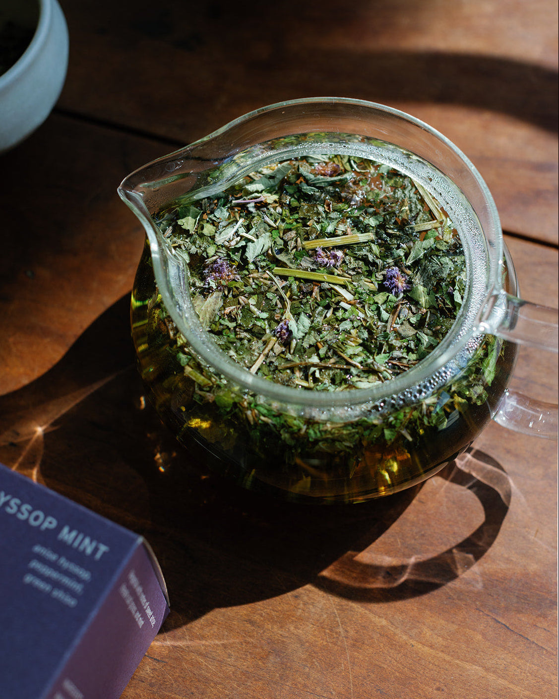 Looseleaf herbal tea in a glass teapot on a wooden surface with a box labeled 'Hyssop Mint' in the background.