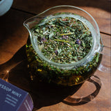 Looseleaf herbal tea in a glass teapot on a wooden surface with a box labeled 'Hyssop Mint' in the background.