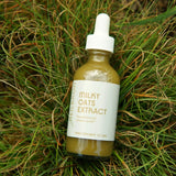 A clear dropper bottle of cloudy and green milky oats extract resting in green grass