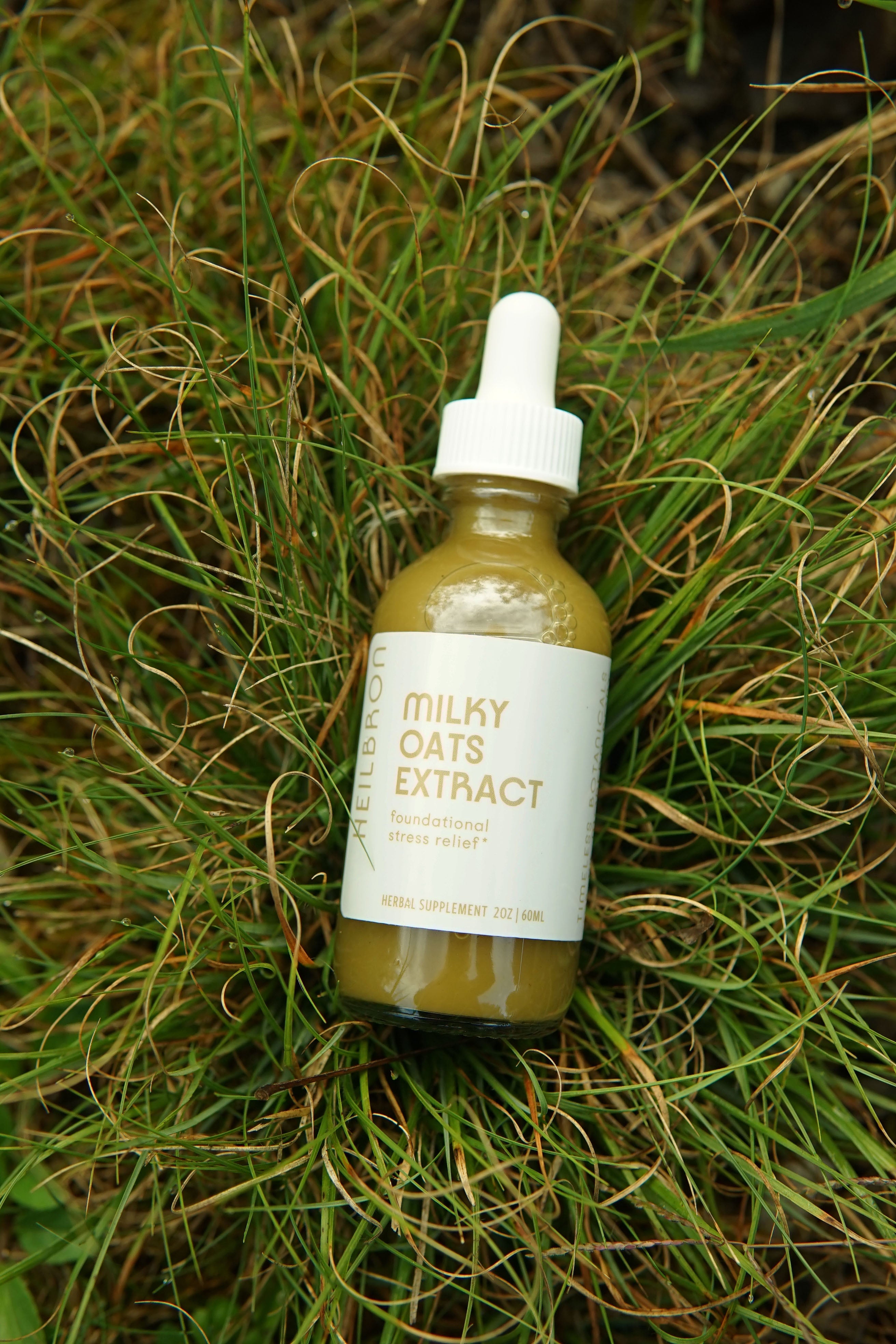 A clear dropper bottle of cloudy and green milky oats extract resting in green grass